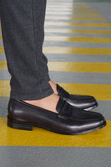 Black leather loafers paired with dark textured trousers on a striped yellow-gray floor.