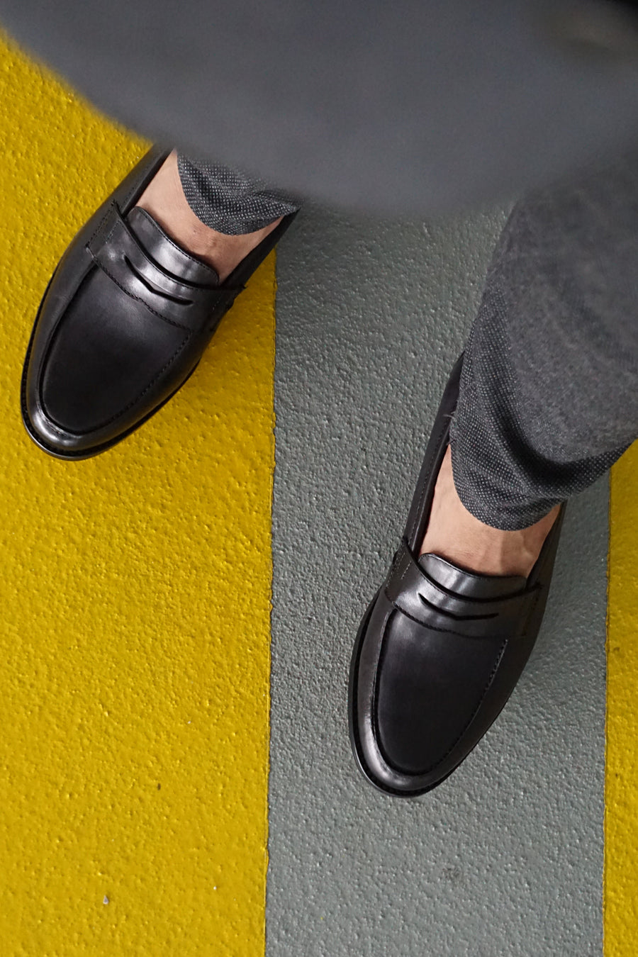 Black leather loafers paired with gray trousers on a yellow-gray striped floor