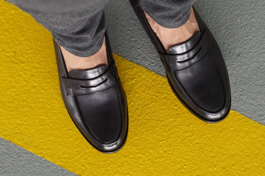 Black leather loafers on yellow and gray textured floor, paired with gray pants.